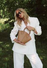 Ruda odinė rankinė su struktūrizuotu dizainu, dvigubais trumpais rankenomis ir matomais siuvimo detalėmis. Dėvima su baltu kostiumu žalumos fone.