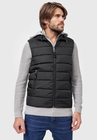 Sort puffer vest med vandret søm, lynlås-lommer og høj krave. Laget over en lysegrå trøje med ribbede manchetter.