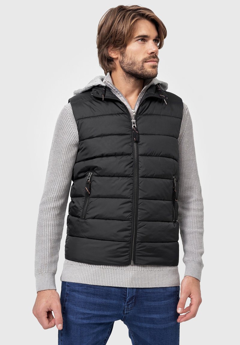 Sort puffer vest med vandret søm, lynlås-lommer og høj krave. Laget over en lysegrå trøje med ribbede manchetter.