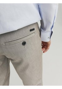 Světlé šedé kalhoty na míru s hladkou texturou, zapínání na knoflík na zadní kapse a jemným tkaným vzorem. Viditelná košile v jemných modrých tónech.