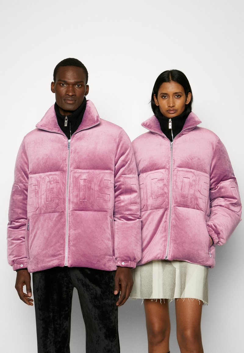 Giubbotti puffer in velluto rosa con colli alti, zip frontali e dettagli del logo in rilievo. Indossati da due persone su uno sfondo neutro.
