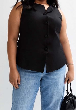 Frau trägt ärmellose schwarze Weste mit Knöpfen und hellblaue Jeans, hält eine schwarze Lederbeuteltasche in der rechten Hand.