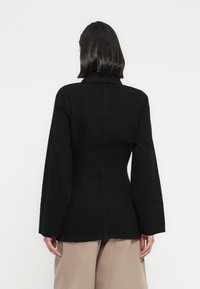 Μαύρο τζιν μπο υφάν με δομημένο σχεδιασμό, γιακά και μακριά, φαρδιά μανίκια. Διαθέτει ορατές ραφές και εφαρμοστή σιλουέτα.