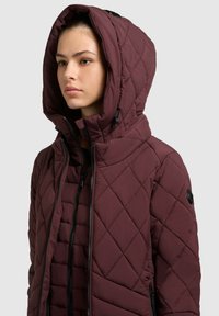 Tânără purtând o geacă burgundă matlasată cu glugă, cu două fermoare negre și buzunare laterale, orientată spre stânga, pe un fundal gri.