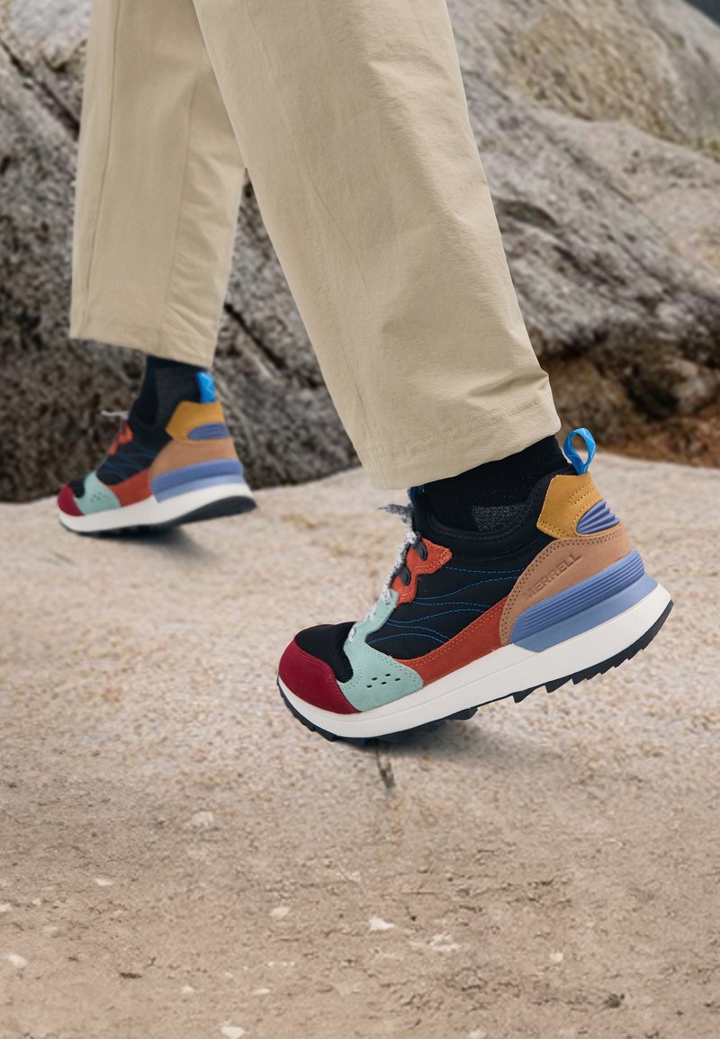 Farverig sneaker med varierede materialer: sort mesh, flerfarvet ruskind, blå accenter og en kraftig hvid sål med tekstureret mønster.