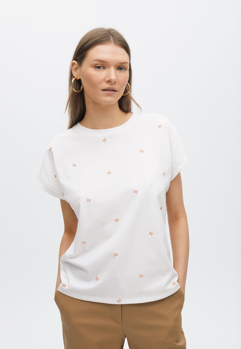 T-shirt blanc à manches courtes avec une texture lisse, présentant de petits logos "M" orange dispersés. Associé à un pantalon marron clair.