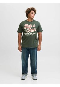 Olivengroen katoenen T-shirt met witte en rode tekst, gedragen met blauwe denim jeans en grijze sneakers. Eenvoudig ontwerp, losse pasvorm.