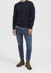 Marinblå kabelstickad tröja med rund halsringning, kombinerad med ljusblå denimjeans och bruna mocka Chelsea-boots. Klassisk avslappnad klädsel.