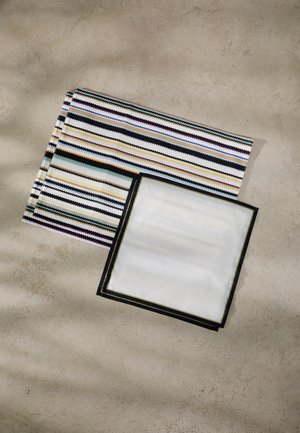 Striped textile placemat in multicolors, featuring zigzag patterns. Next to it, a white placemat bordered in black. Textured surfaces visible.