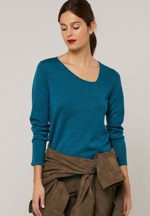 Vrouw met donker haar, gekleed in een teal gekleurde top met lange mouwen en een bruine jas gestrikt rond haar taille, die naar beneden kijkt met rode lippenstift.