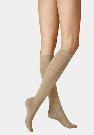 Legs crossed wearing beige knee-high compression socks made of smooth, lightweight fabric against a plain background.