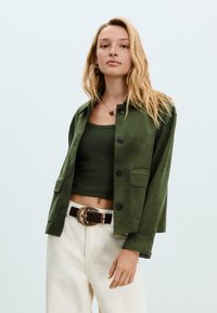 Giacca in suede verde con chiusura a bottoni e due tasche frontali, indossata sopra un top smanicato verde scuro. Abbinata a pantaloni color crema e una cintura decorata.