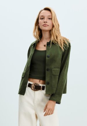 Veste en daim vert avec fermeture à bouton et deux poches avant, portée par-dessus un débardeur vert foncé. Assortie avec un pantalon crème et une ceinture décorée.