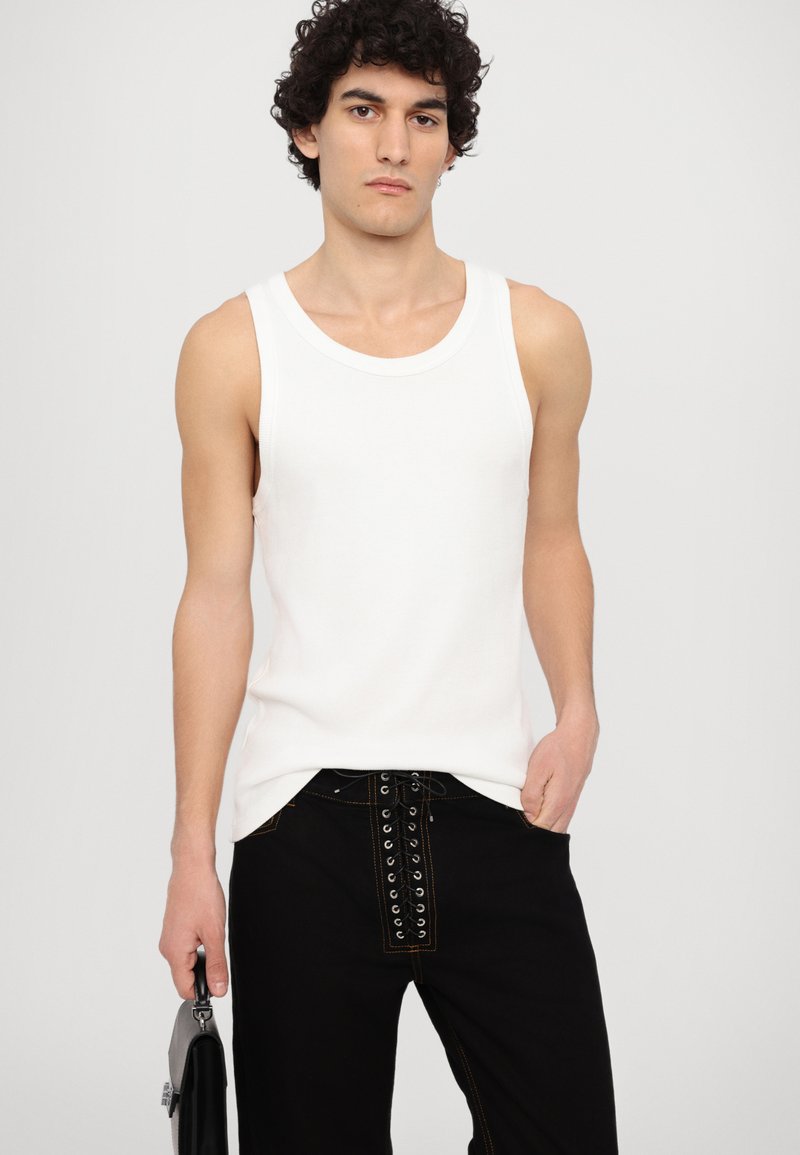 White tank top in ribbed texture paired with black jeans featuring a lace-up front design. Model holding a small black bag.