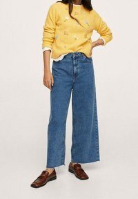 Pull en maille jaune avec broderie florale, jean bleu taille haute à jambes larges, et mocassins en cuir marron avec une finition texturée.