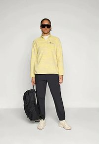 Gul fleece-pullover med halv dragkedja, logotyp på bröstet; svarta byxor, beige sneakers och svart ryggsäck kompletterar outfiten.