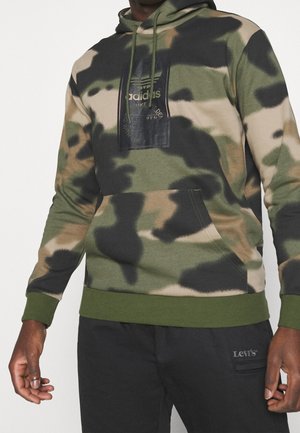 Mężczyzna w zielonej, beżowej i czarnej bluzie z kapturem Adidas w kamuflażu z przednią kieszenią oraz czarnych spodniach Levi's widocznych od talii do nadgarstków.