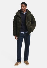 Chaqueta de pana acolchada en verde oscuro con capucha, botones de presión plateados, sudadera azul marino debajo, camisa a cuadros azul claro, jeans oscuros, zapatillas marrones.