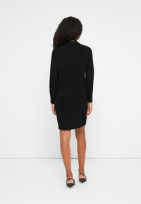 Donna con capelli ricci lunghi, in piedi di spalle, indossa un vestito nero a maniche lunghe fino al ginocchio e scarpe basse nere su uno sfondo bianco.