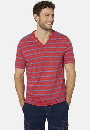 Mann lächelt, trägt rotes Henley-Shirt mit blauen horizontalen Streifen und dunkelblaue Cargohosen, Hände in den Taschen.