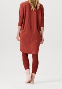 Langes, locker sitzendes Kleid in rostfarben mit dreiviertel Ärmeln, kombiniert mit passenden Leggings und rosa gerippten Socken. Glatte Stofftextur.