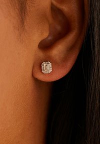 Quadratischer Ohrstecker mit einem klaren, smaragdgeschliffenen mittleren Stein, umgeben von einem Kranz kleiner weißer Steine, gefasst in glänzendem Metall.