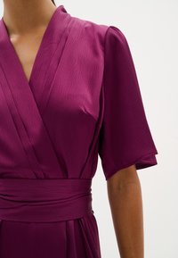 Fuchsia omlottklänning med korta ärmar, texturerad tyg och matchande midjebälte. Har en djup V-ringning och mjuk, flödande fall.