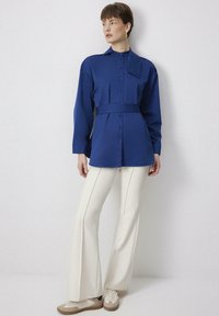 Camicia blu con cintura e maniche lunghe, chiusura a bottoni, abbinata a pantaloni crema a campana e sneakers beige. Tessuto dalla trama liscia.