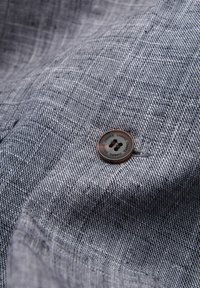Gros plan d’un tissu gris texturé avec un seul bouton foncé cousu, présentant le mot "OMBRE" répété autour de son bord.