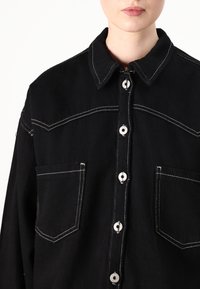 Camisa de denim preta com costura branca, fecho de botão, colarinho e dois bolsos frontais. Tecido texturizado e design clássico.