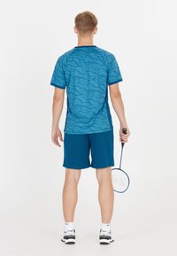 Manlig idrottare klädd i blåmönstrad sportbekädnad håller en badmintonracket och står vänd bakåt mot en vit bakgrund.