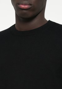 Μαύρο πλεκτό πουλόβερ με στρογγυλή λαιμόκοψη, με απαλή υφή και εφαρμοστή σχεδίαση. Κοντινή λήψη του λαιμού και του άνω κορμού.