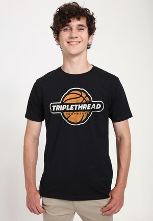 Giovane uomo sorridente, indossa una maglietta nera con un pallacanestro arancione e il logo "TRIPLETHREAD HOOPWEAR", pantaloni beige, in piedi davanti a uno sfondo semplice.