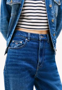 Tmavě modré džínové kalhoty s klasickým pětikomorovým designem, malým logem na přední kapse a vysokým pasem. Pod nimi proužkované tričko.