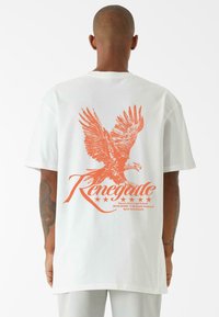 Baltas T-krekls ar lielu oranžu ērgļa grafiku un vārdu "Renegade" ar zvaigznēm zem tā. Īsas piedurknes un brīvs piegriezums.