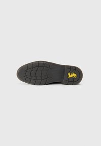 Suela de zapato negra con profundas estrías y un logo de león amarillo cerca del talón sobre un fondo claro y liso.