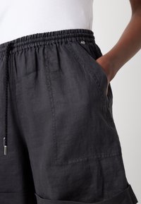 Personne portant un short en lin noir avec une taille élastique et un cordon de serrage, une main dans la poche latérale, sur un fond clair uni.
