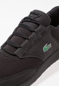 Baskets Lacoste noires en tissu texturé, avec des détails en daim, des lacets noirs et un logo crocodile vert sur le côté.