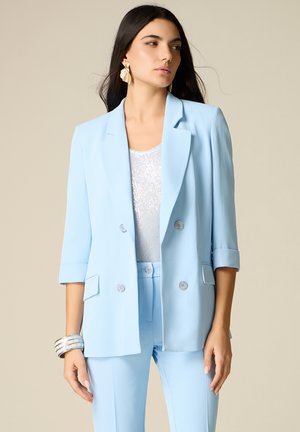 Femme aux longs cheveux foncés portant un blazer bleu clair et un pantalon assorti, un haut blanc texturé, des boucles d'oreilles en forme de coquillage doré et des bracelets argentés.