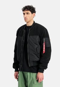 Muž oblečený v černé fleecové bundě na zip a nylonové bundě s červeným štítkem "Odstranit před letem" na rukávu, v kombinaci se zelenými cargo kalhotami.