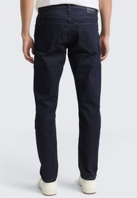 Ciemnoniebieskie dżinsy o dopasowanym kroju, z dwoma tylnymi kieszeniami i subtelną naszywką z logo w pasie. Gładka tekstura, standardowa długość.