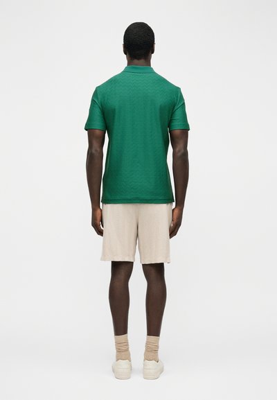 Άνδρας που στέκεται πλάτη, φορώντας πράσινο πόλο με υφή και κοντά μανίκια, μπεζ σορτς, μπεζ κάλτσες και λευκά αθλητικά παπούτσια, σε απλό φόντο.