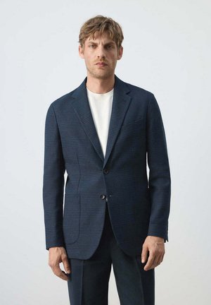 Mann trägt einen dunkelblau karierten Blazer über einem weißen Hemd, steht vor einem einfarbigen hellgrauen Hintergrund.