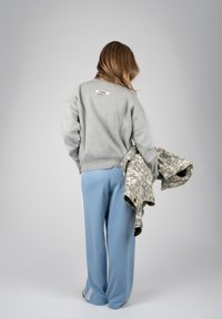 Femme de dos portant un pull gris et un pantalon bleu, tenant une veste camouflage dans la main gauche sur un fond uni.