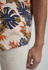 Camicia a maniche corte realizzata in tessuto leggero, caratterizzata da un motivo di foglie tropicali in tonalità di blu navy, arancione e beige. L'orlo è leggermente curvo.