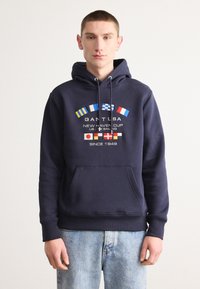 Felpa blu navy con tasca a marsupio, decorata con diverse bandiere nautiche internazionali e testo. Realizzata in tessuto morbido, con cappuccio regolabile.