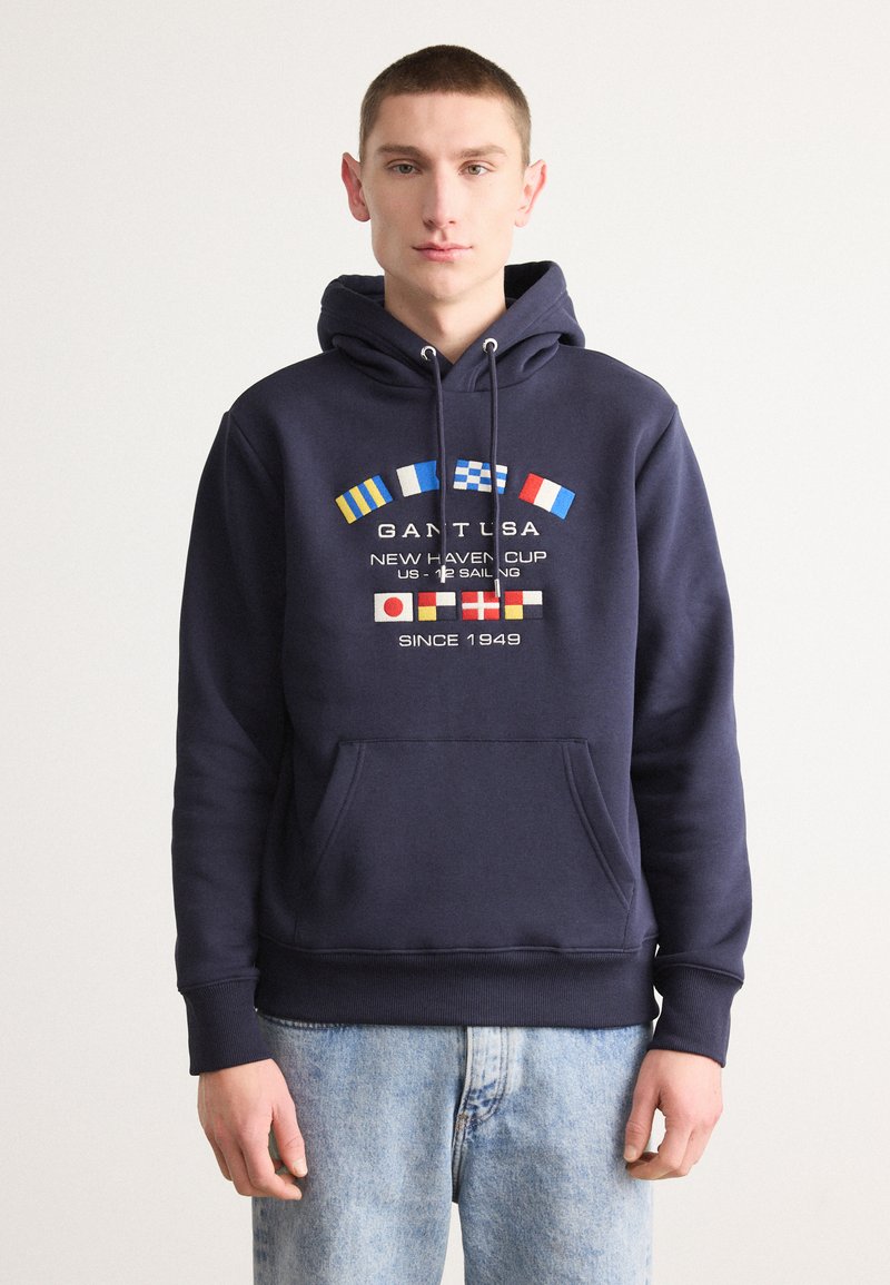 Felpa blu navy con tasca a marsupio, decorata con diverse bandiere nautiche internazionali e testo. Realizzata in tessuto morbido, con cappuccio regolabile.
