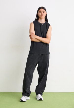 Top nero senza maniche abbinato a jeans larghi grigi scuri. Il modello indossa sneakers bianche e una sottile catena d'argento. Lo sfondo è minimalista.