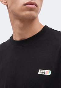 Κοντινό του κάτω μέρους προσώπου και του λαιμού ενός ατόμου που φοράει μαύρο T-shirt με μικρό λευκό λογότυπο που γράφει "JUUE" και τρεις πολύχρωμες ρίγες.