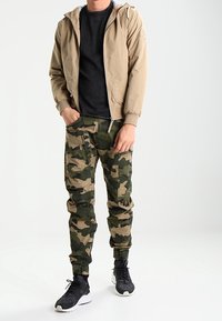 Chaqueta beige con cremallera y capucha sobre una camiseta negra, combinada con pantalones cargo de camuflaje y zapatillas negras. Cuenta con puños elásticos y bolsillos.